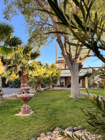 A well-maintained garden with a stone fountain, palm trees, and a building with a tiled roof.