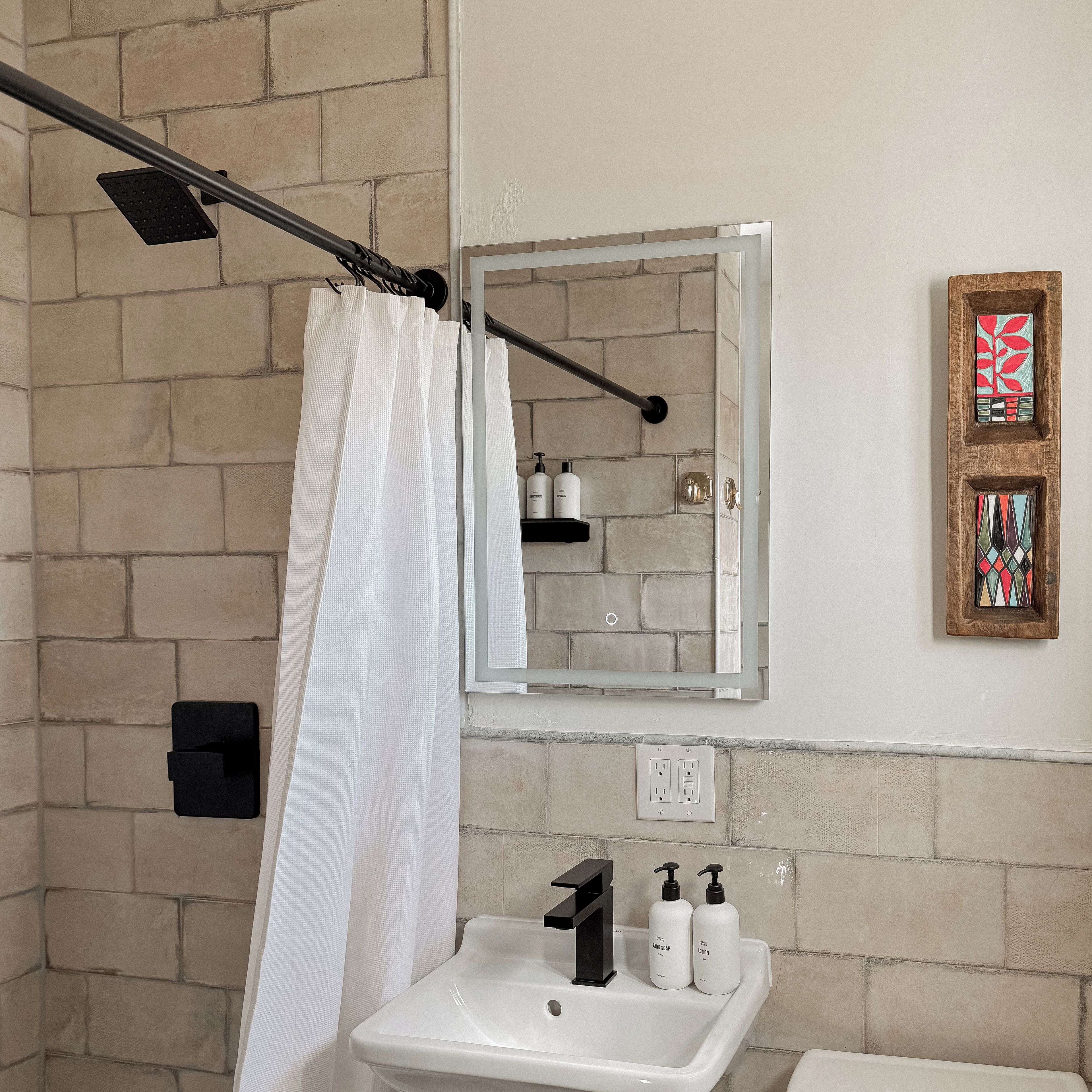 A bathroom with a shower curtain, a mirror, a sink with two bottles of soap, and a wall-mounted shelf with toiletries.