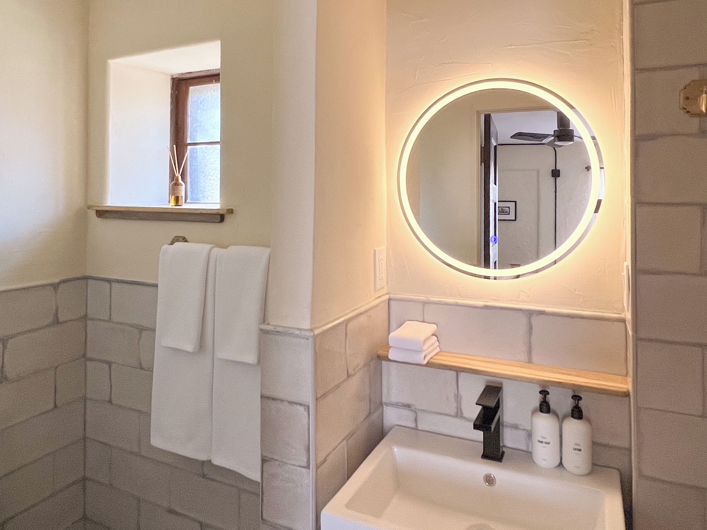 Un baño con un lavamanos blanco, un espejo con luz, un perchero para toallas y una ventana.