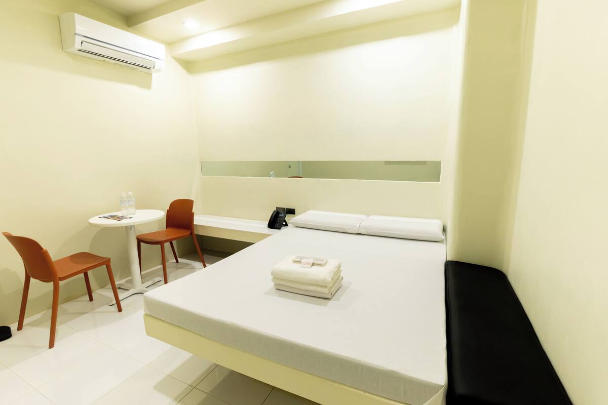 A hotel room with a bed, a desk, two chairs, a small table with a bottle, and an air conditioning unit.