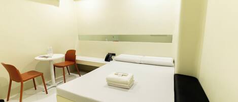 A hotel room with a bed, a desk, two chairs, a small table with a bottle, and an air conditioning unit.
