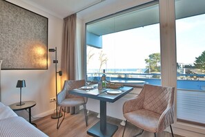 A modern hotel room with a large window offering a view of the sea and a balcony. There is a dining table with chairs, a lamp, and a bed with a view.