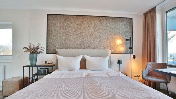 Ein modernes Hotelzimmer mit einem großen Bett, einem Nachttisch mit Radio, einer Vase mit Trockenblumen, einem Sessel und Blick ins Freie.