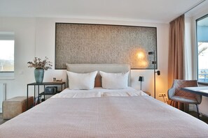 Una habitación de hotel moderna con una cama grande, una mesita de noche con una radio, un jarrón con flores secas, una silla y vistas al exterior.