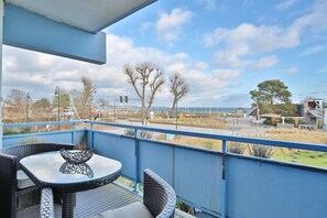 Varanda com mobiliário de vime e mesa de vidro, com vista para uma rua com semáforos e uma praia ao longe.