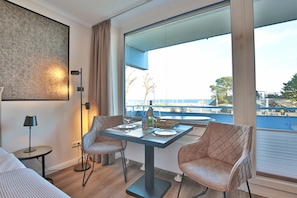 A modern hotel room with a large window offering a view of the sea and a balcony. There is a dining table with chairs, a lamp, and a bed with a view.