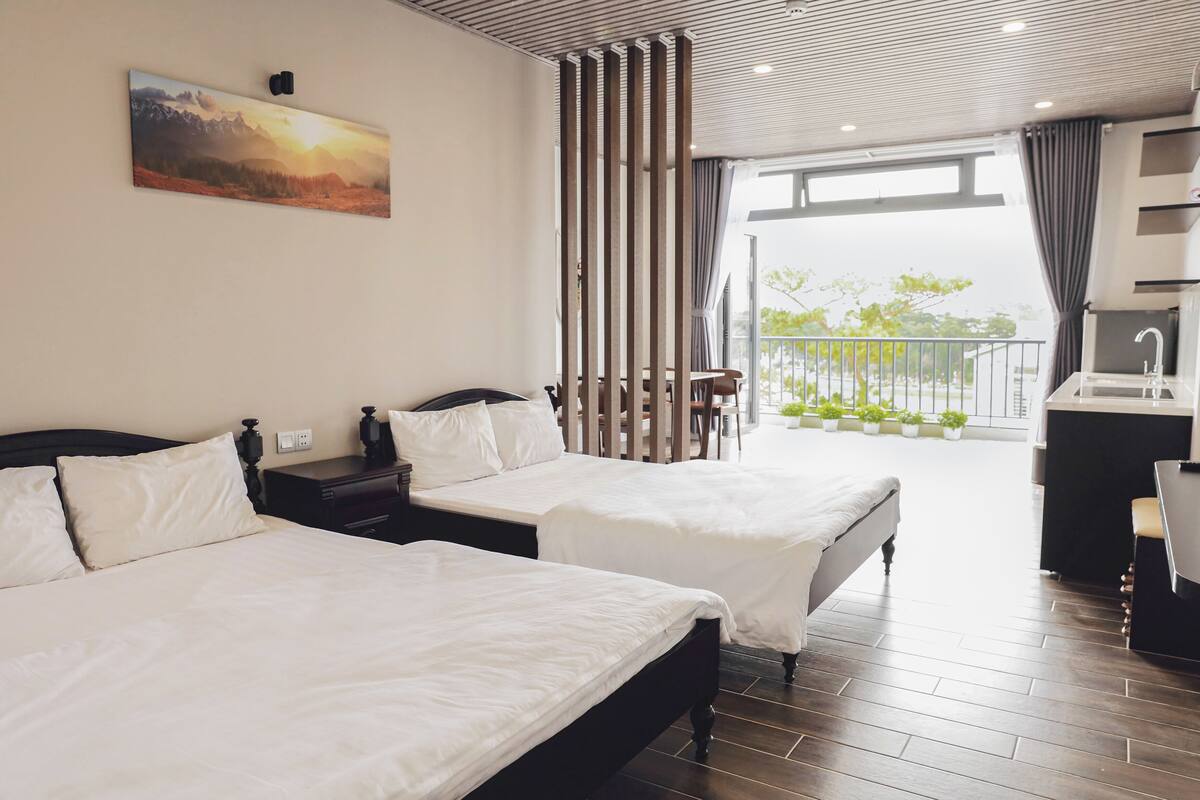 A hotel room with two beds, a large window, a balcony with plants, and a painting on the wall.