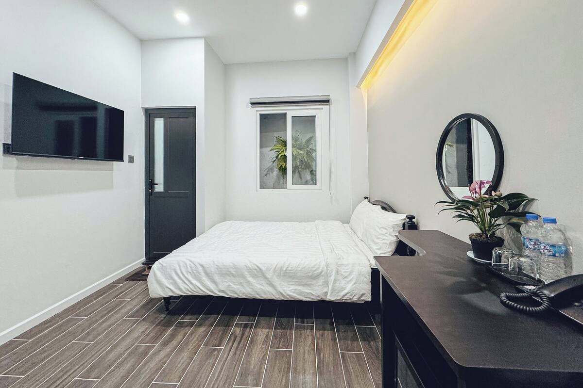A modern bedroom with a bed, a flat-screen TV mounted on the wall, a small window, a bedside table with a mirror, and a telephone.