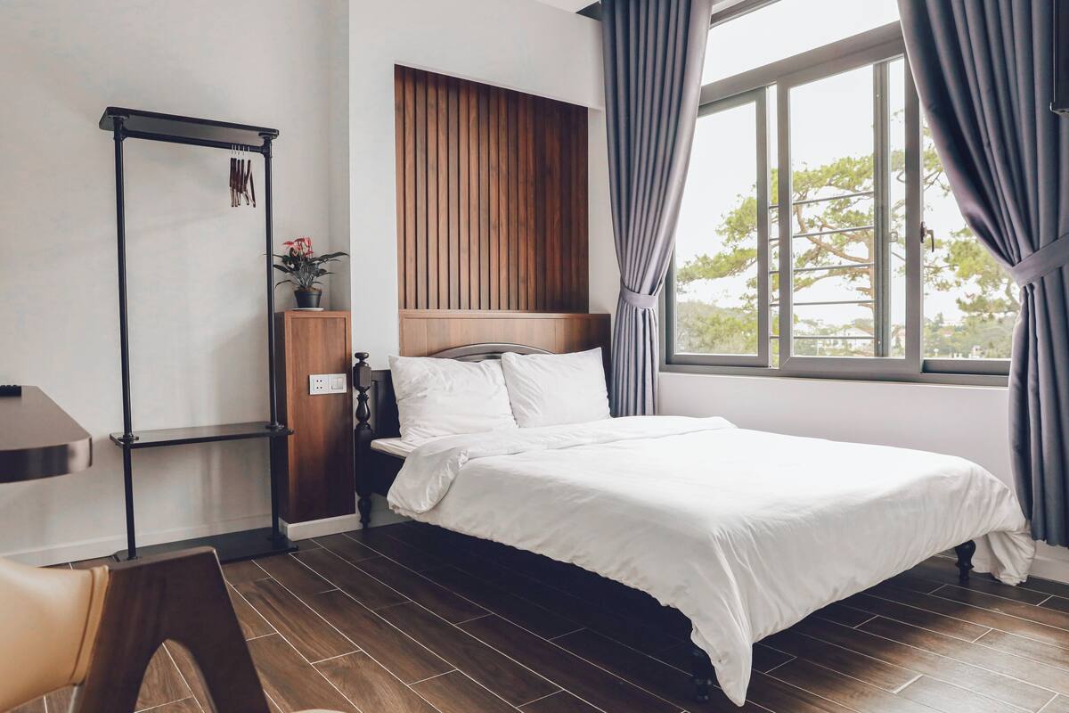 A neatly arranged bedroom with a bed, a wooden headboard, a window with curtains, and a small table with a plant.
