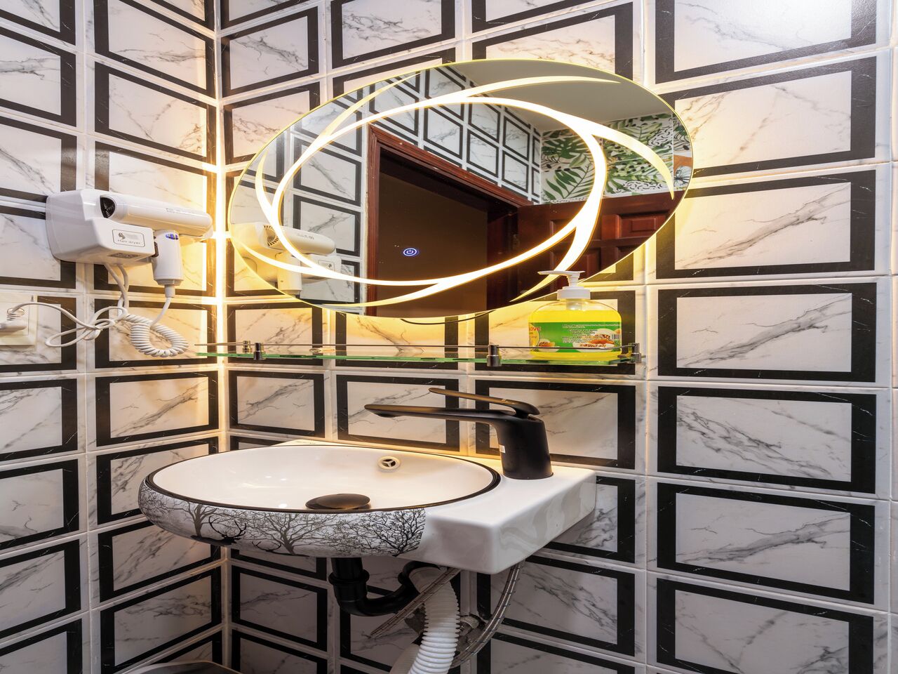 Een badkamer met een marmerlook wand, een ronde spiegel met een rondomgaande verlichting, een wastafel met een ontwerp van een boom, en een handdoekdispenser.