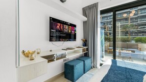 Een moderne woonkamer met een flatscreen televisie aan de muur, een inbouwkast, een zithoek met een blauwe poef en een groot raam met uitzicht op het buitenland.