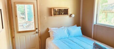 A bedroom with a bed, a door, a window, and a wall-mounted light fixture.