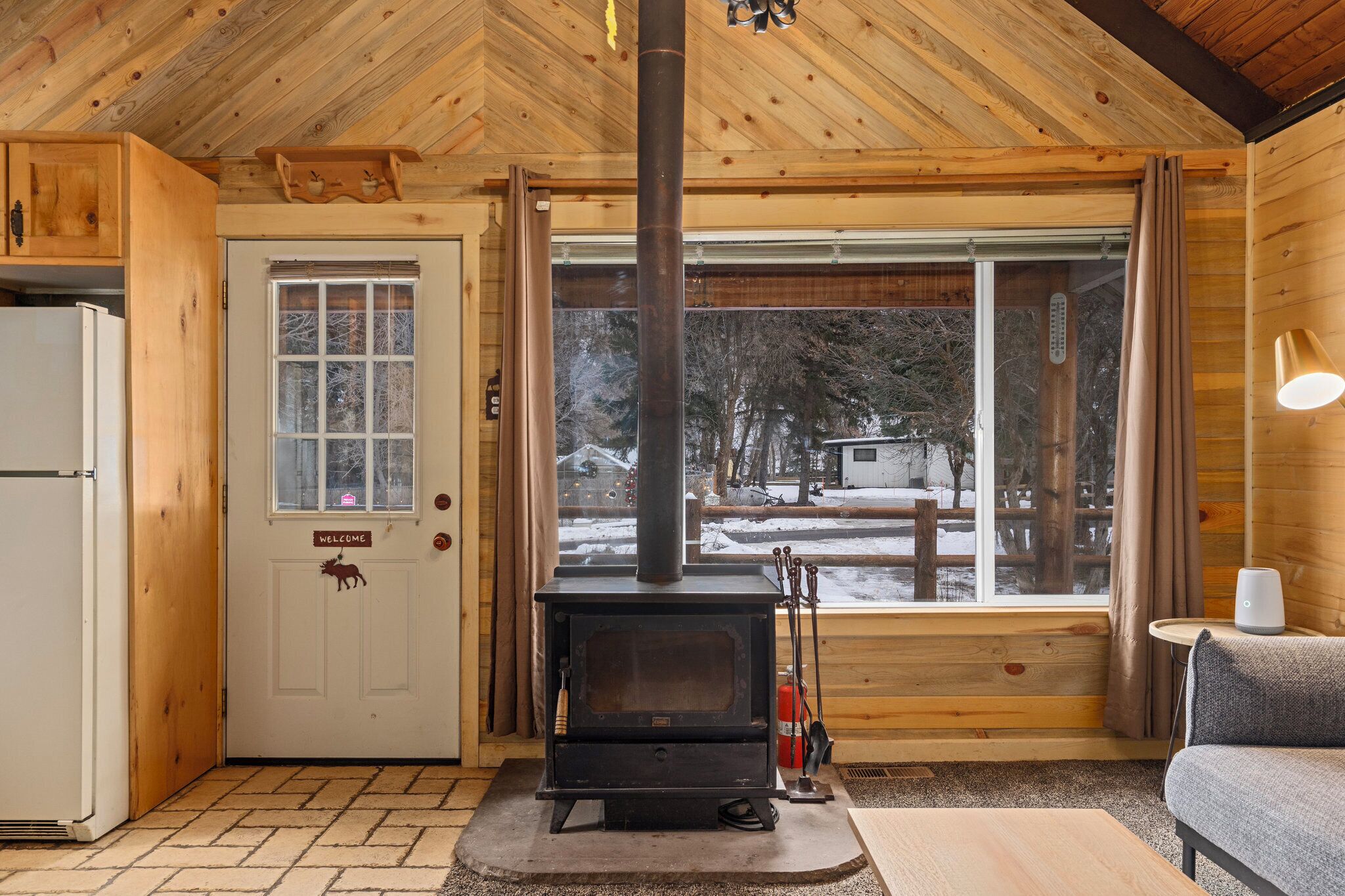 L’intérieur d’un chalet en bois, avec un poêle à bois, une porte blanche ornée d’une pancarte de bienvenue et une vue sur un paysage enneigé, visible à travers de grandes fenêtres.