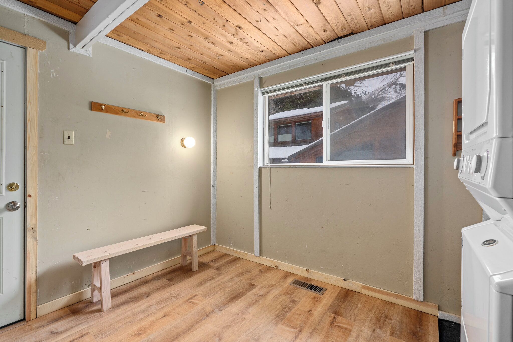 Une pièce avec un banc en bois, une fenêtre donnant sur un paysage enneigé, une porte blanche et un appareil électroménager blanc.