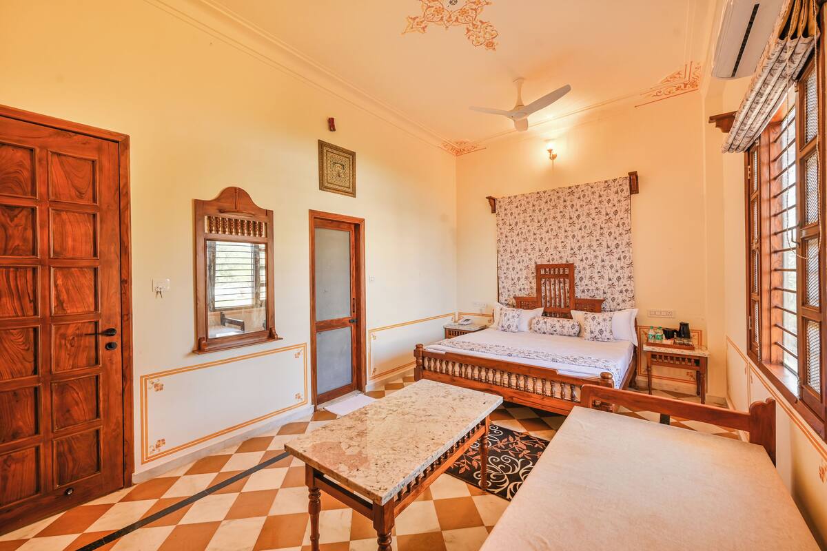 Quarto com cama de madeira, uma mesinha, mesa de cabeceira e janela com cortinas estampadas.
