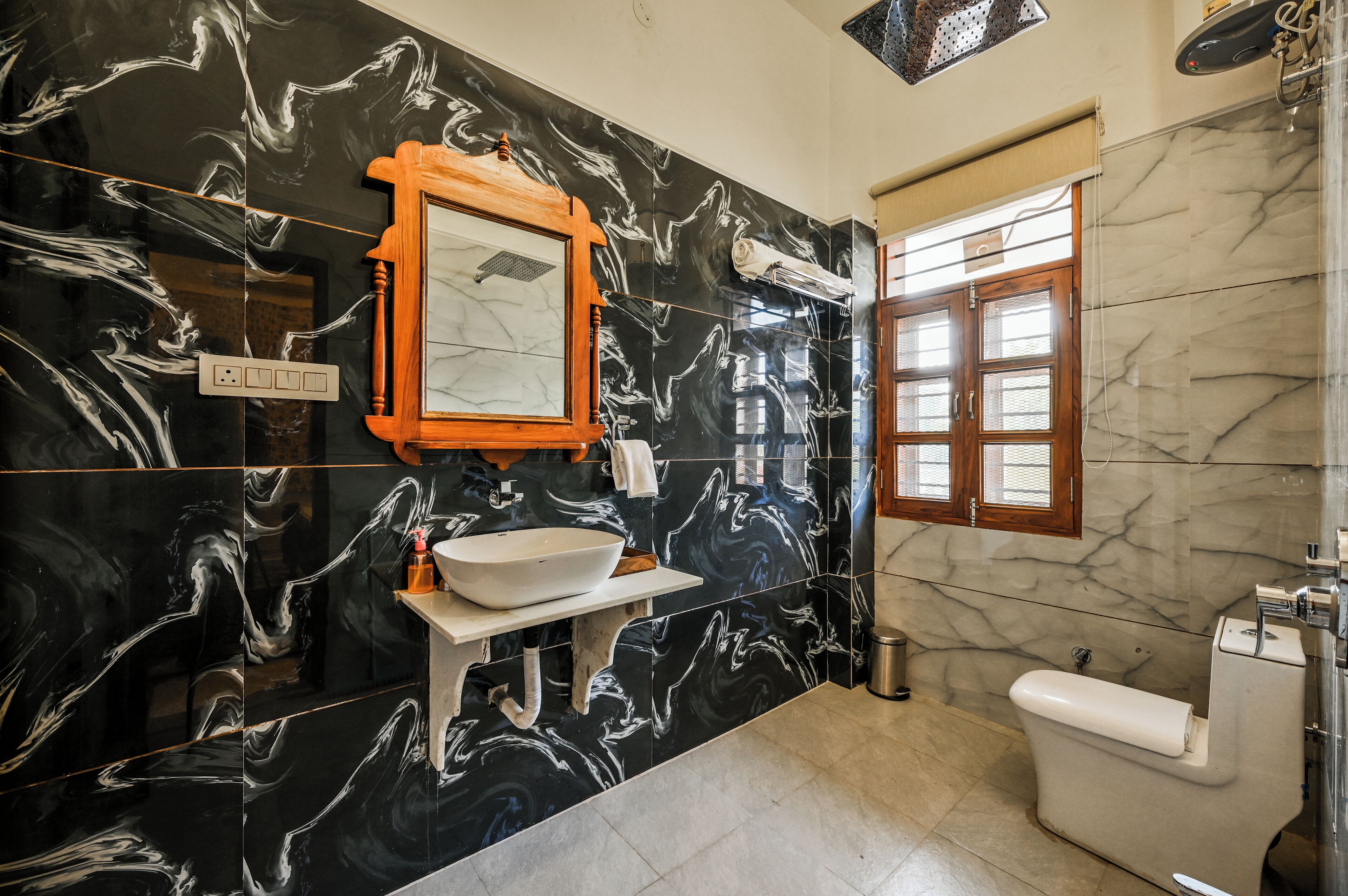 Un baño con pared de mármol negro, espejo con marco de madera, lavabo blanco y un inodoro.