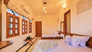 A hotel room with two beds, wooden furniture, a small table, and a window with a view. - Tryambakam Villa (Bhilwara)