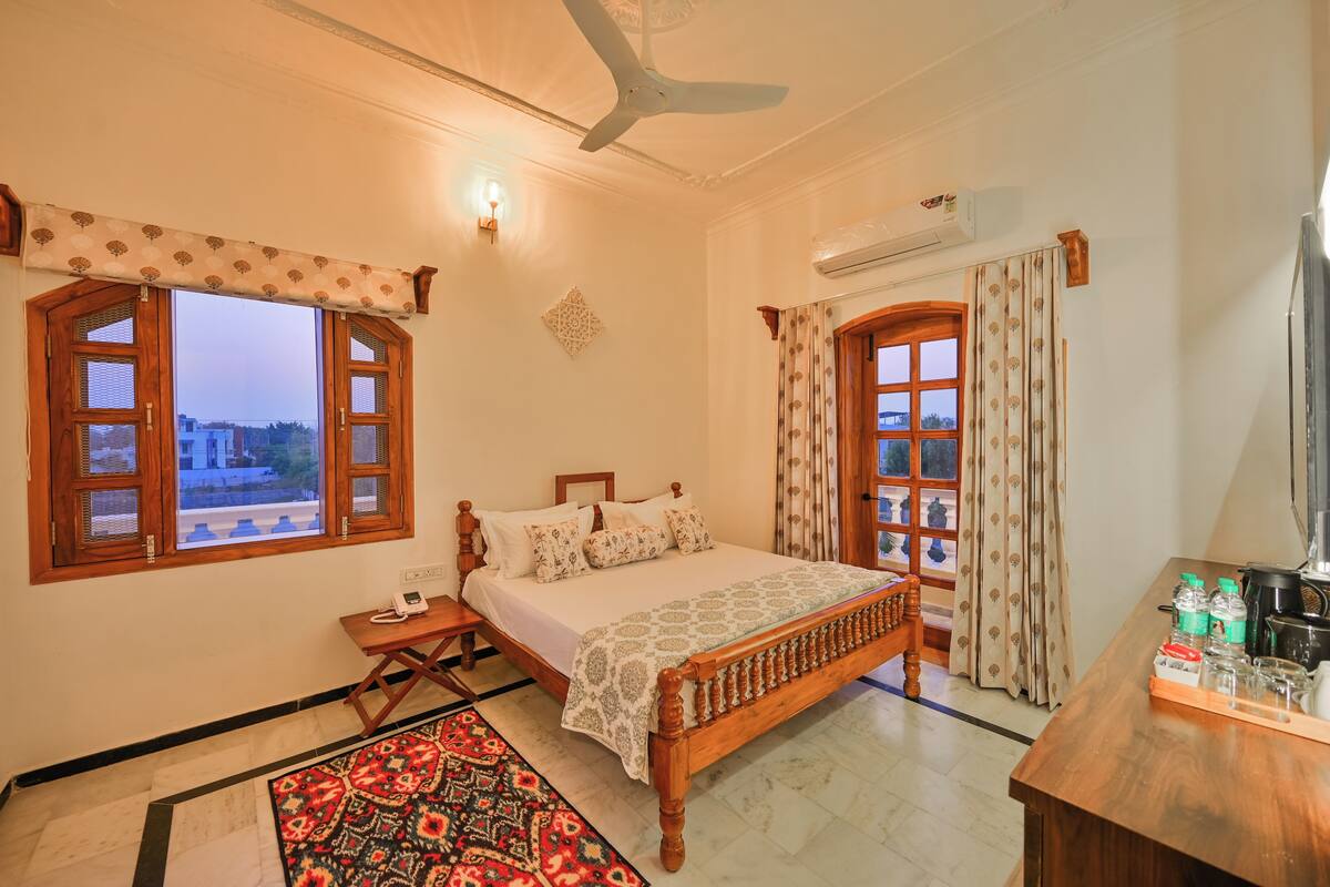 Quarto com cama de madeira, duas janelas com cortinas, ventilador de teto e mesa de cabeceira.
