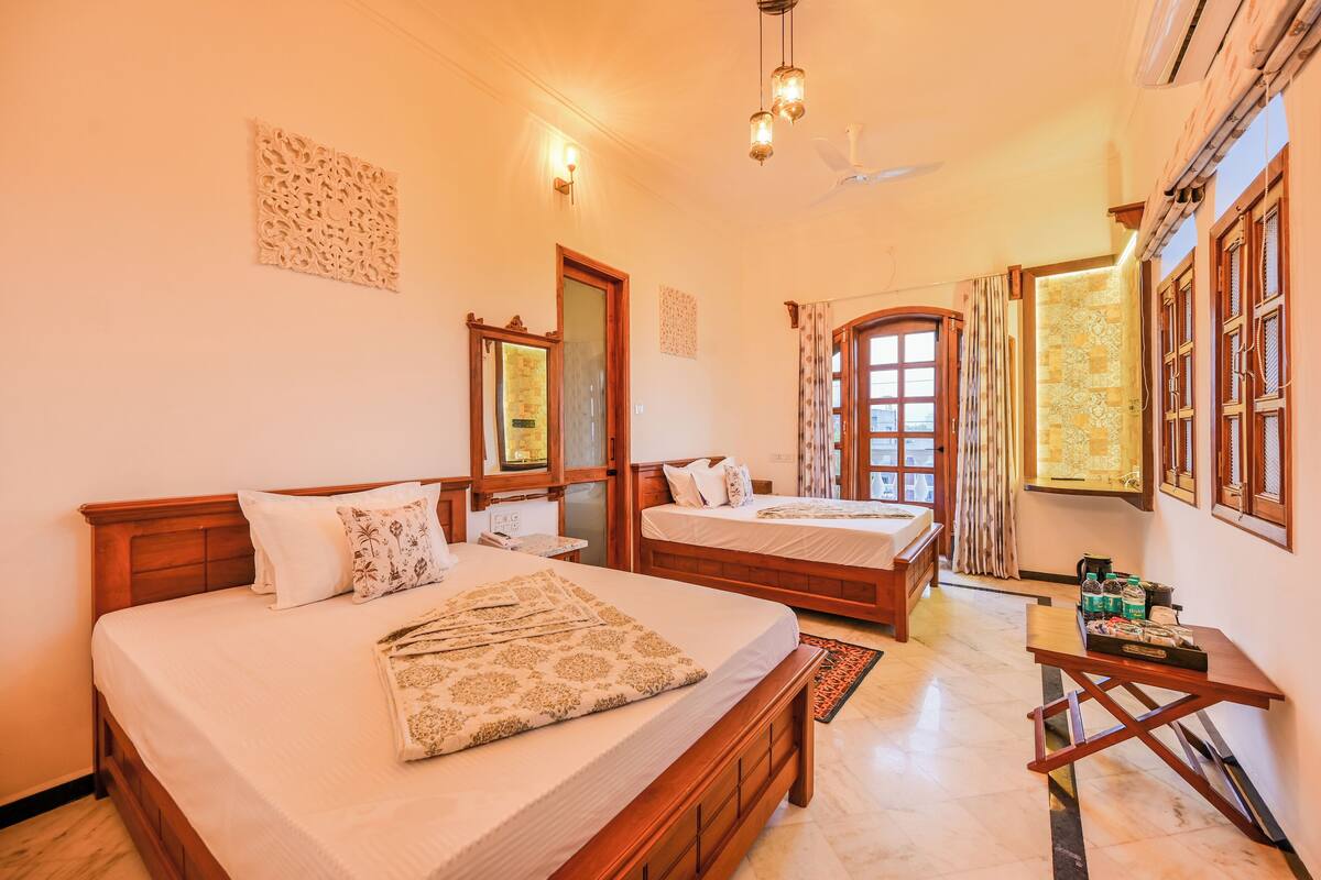 Quarto de hotel com duas camas de madeira, uma mesinha com amenidades, um detalhe decorativo na parede e uma janela com cortinas.