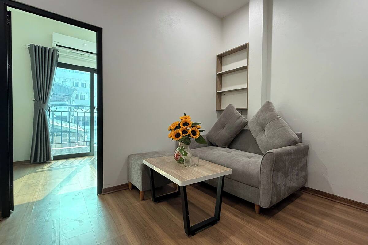 Un salon avec un canapé gris, une petite table en bois ornée d’un vase de tournesols et un balcon avec des rideaux gris.