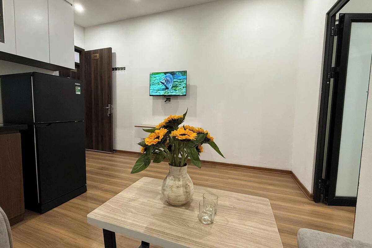 Une cuisine de taille modeste, avec un réfrigérateur noir, un meuble en bois, une table blanche ornée d’un vase de tournesols et un téléviseur fixé au mur.