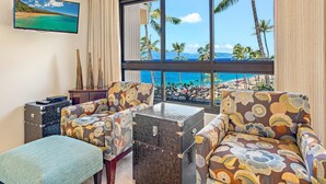 Una habitación con un ventanal grande con vistas a una playa, dos sillones estampados, una mesita y un jarrón decorativo.