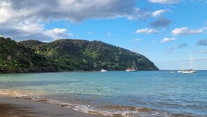 Ein Strand mit ruhigem Wasser, Booten und einem bewaldeten Hügel im Hintergrund.