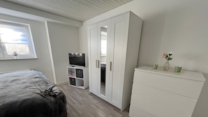 Ein Schlafzimmer mit Bett, Kleiderschrank, Fernsehschrank und einem Fenster mit Jalousien.