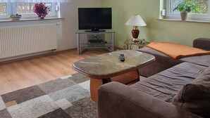 Un salon avec un canapé, une petite table, un téléviseur, une lampe et une fenêtre ornée de plantes.