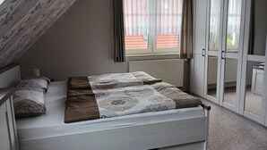Une chambre avec un plafond en pente, un lit recouvert d’une literie à motifs, une fenêtre ornée de rideaux légers, et une vue sur un bâtiment aux toits rouges à l’extérieur.