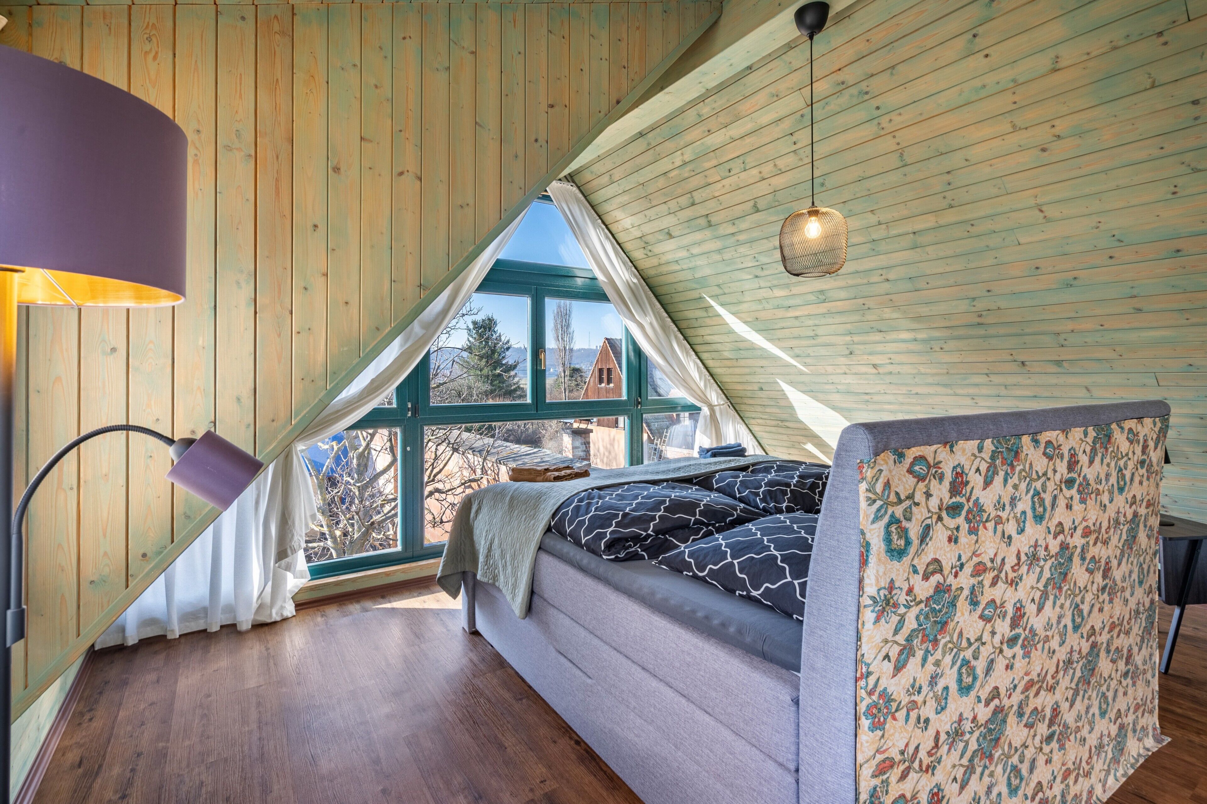 A bedroom with a bed, a lamp, a window, and a view of the outside.