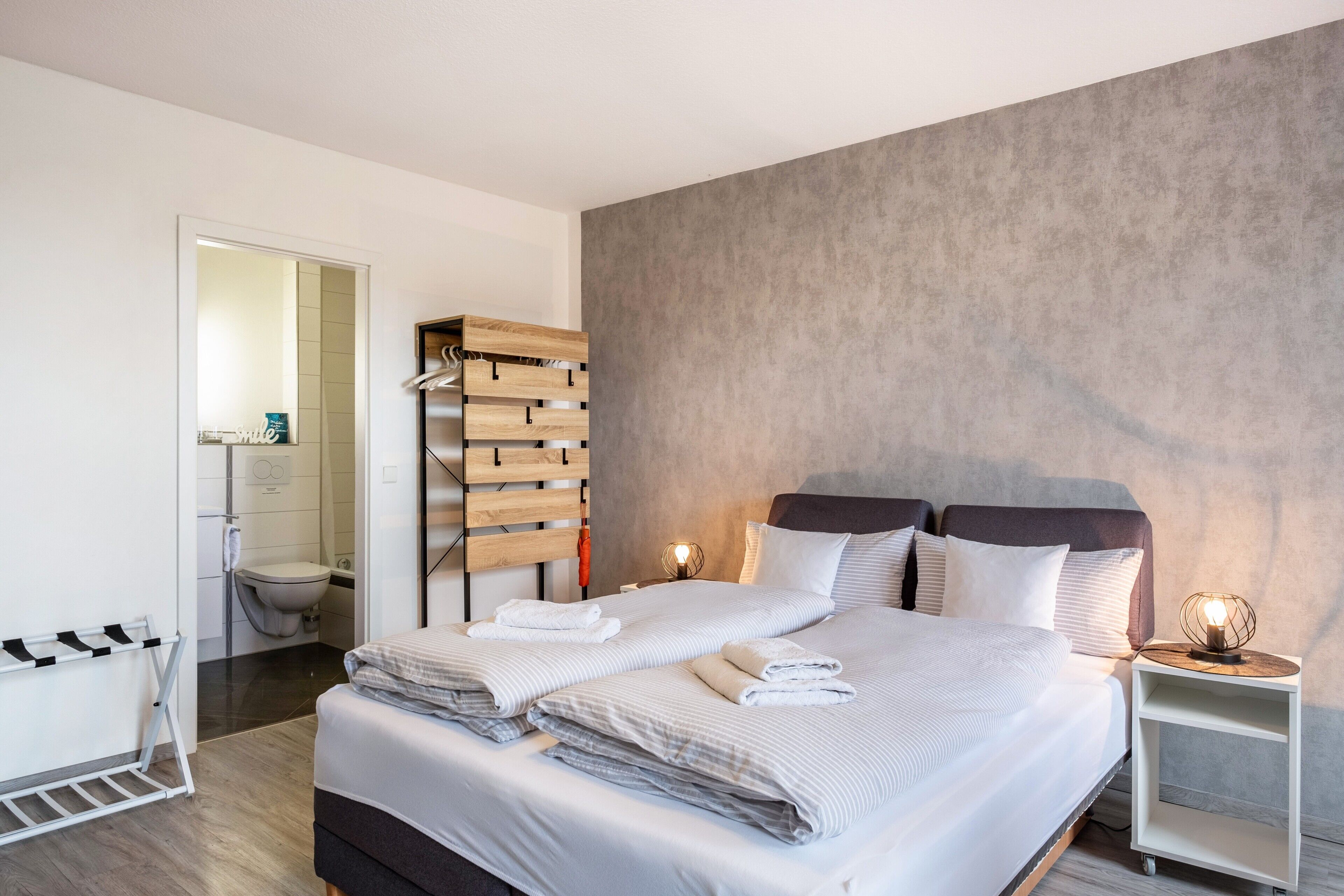 Quarto moderno com uma cama grande, um guarda-roupa de madeira, uma pequena mesa de cabeceira com abajur, e um banheiro visível através de uma porta aberta.