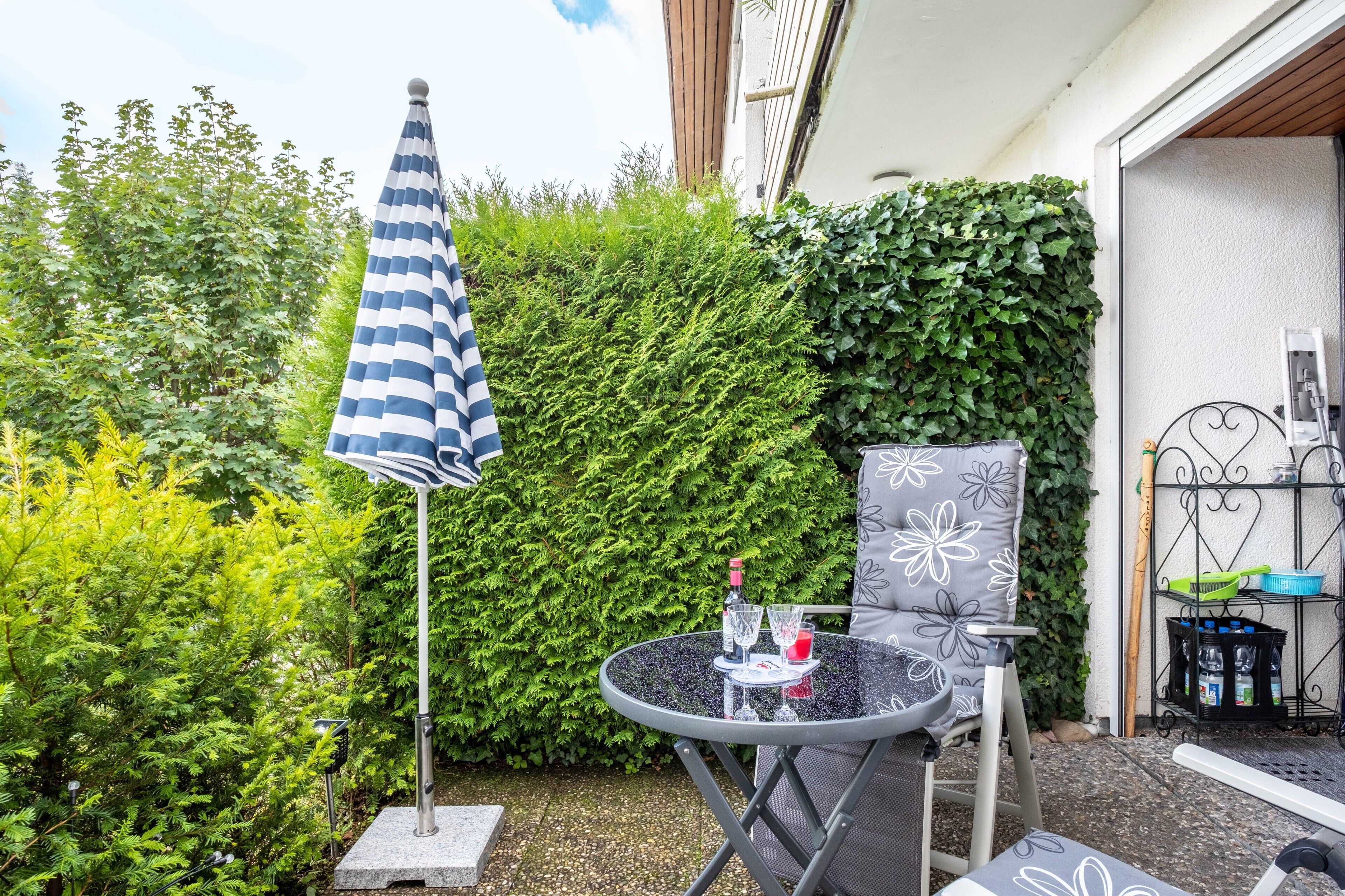 Eine Terrasse im Freien mit einem runden Tisch, einem Stuhl, einem gestreiften Sonnenschirm und einem Heckenabschnitt.