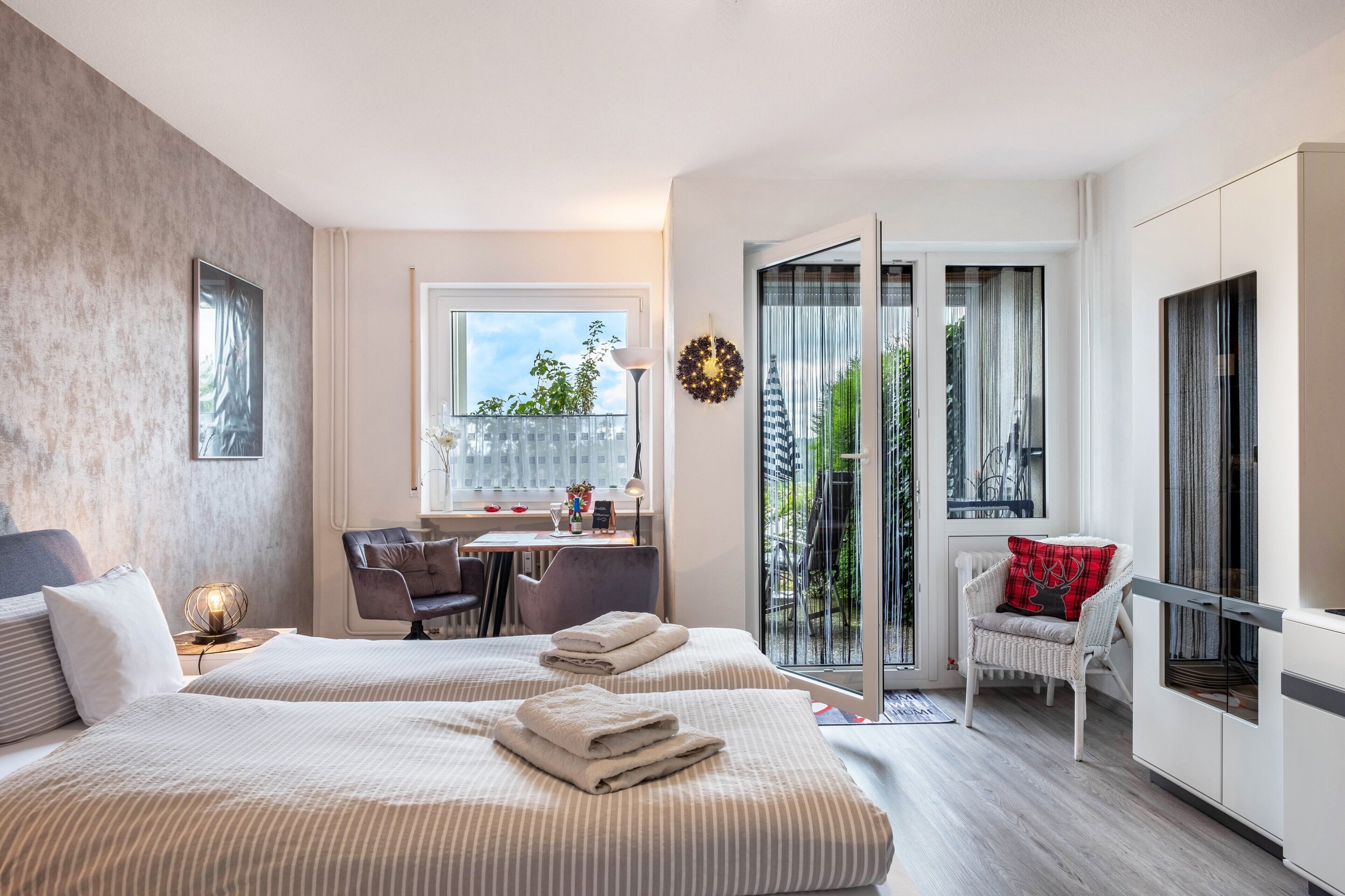 A modern hotel room with two beds, a desk, a chair, a balcony with a view, and a wardrobe.