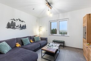 A modern living room with a sofa, a coffee table, a bench, a wall with a mountain and forest decal, and a window with a view.