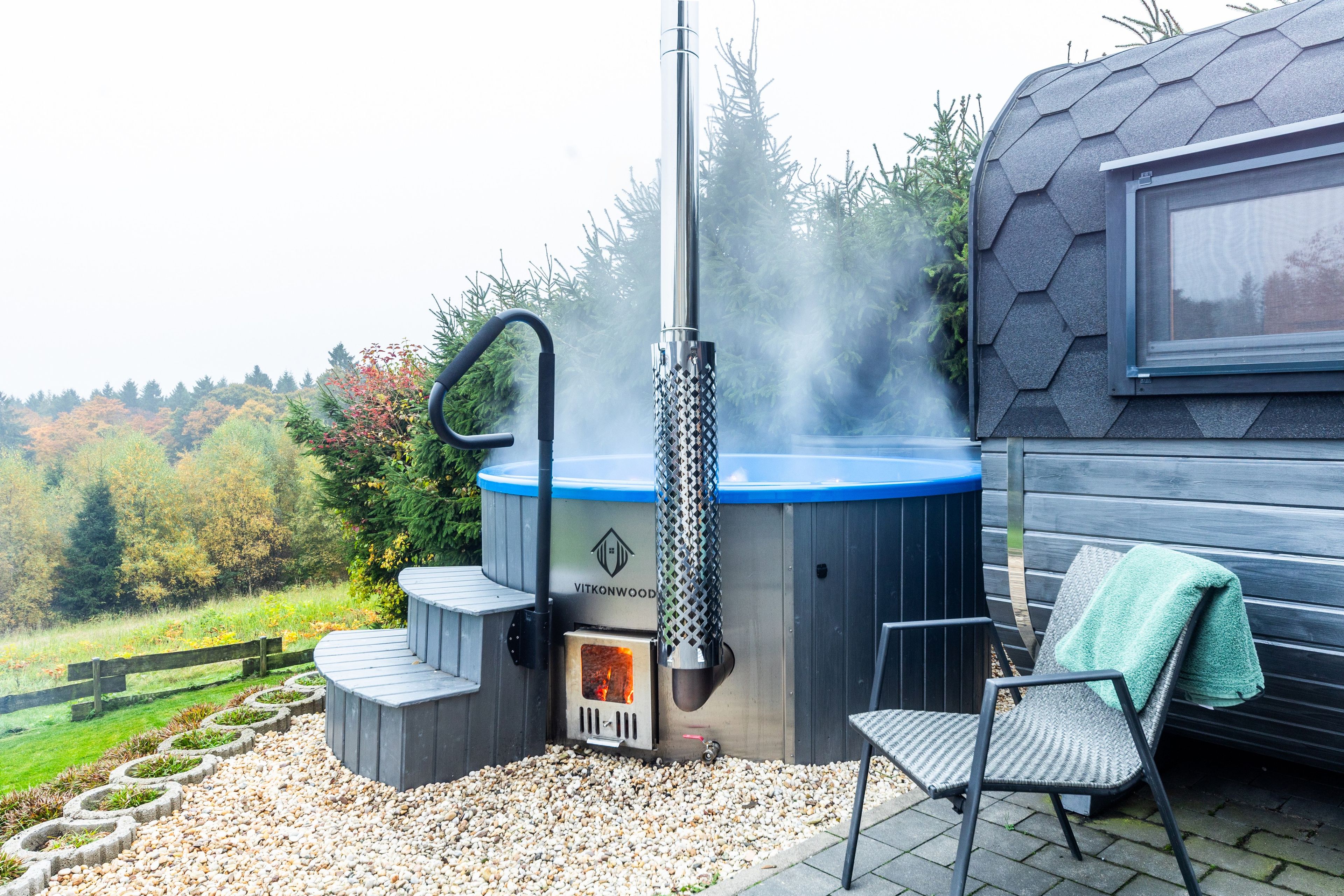 Ein Whirlpool mit Schornstein, umgeben von einer Holzterrasse mit Stuhl und Handtuch, vor einer Kulisse aus Bäumen und einem Holzzaun.