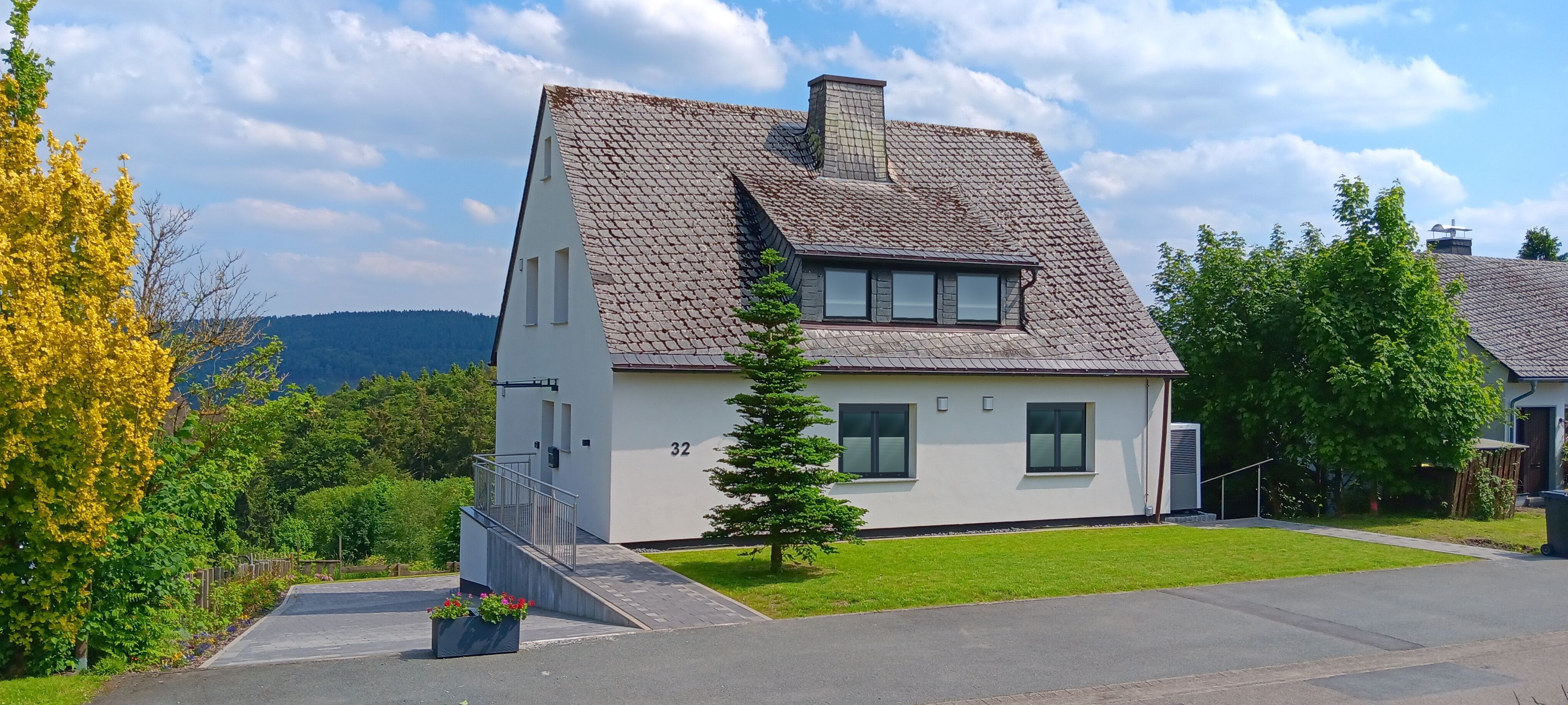 Ein zweistöckiges Haus mit geneigtem Dach, einem kleinen Vorgarten und der Hausnummer 32 an der Seite.