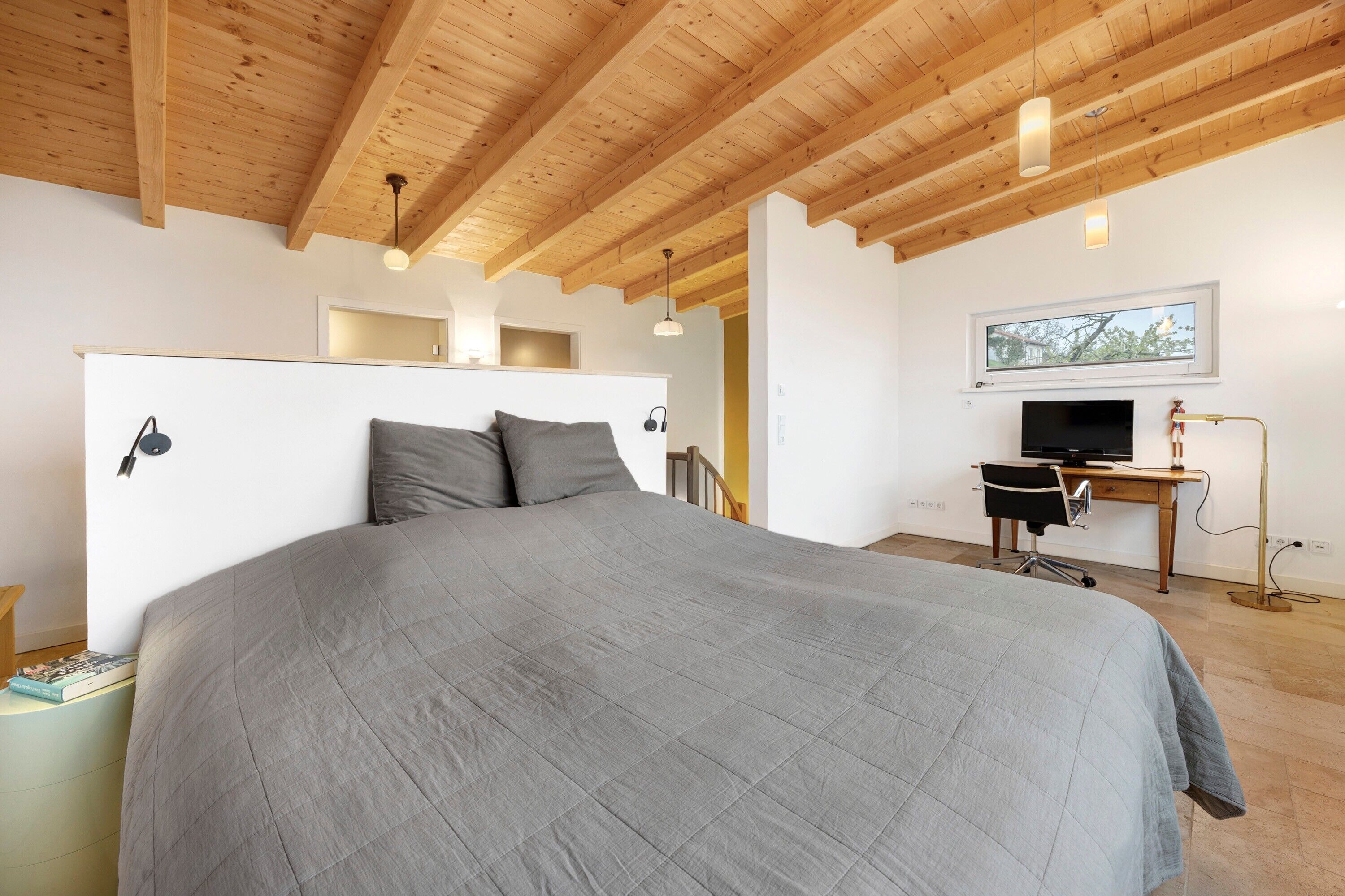 Une chambre avec un grand lit, des poutres apparentes en bois au plafond, un bureau avec un ordinateur et une fenêtre.