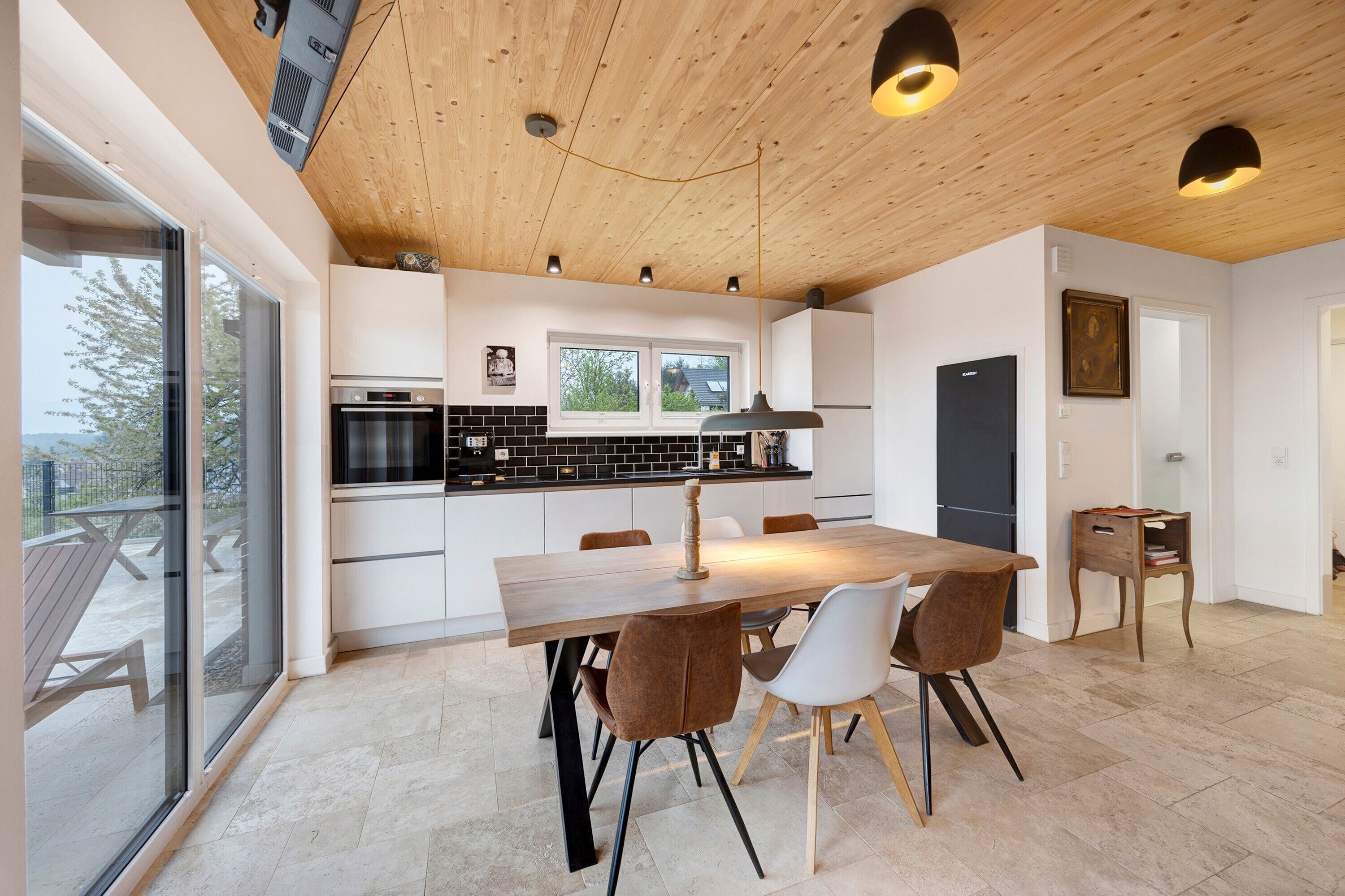 A modern kitchen with a wooden ceiling, a dining area with a large table and chairs, and a view through a glass door to an outdoor patio.