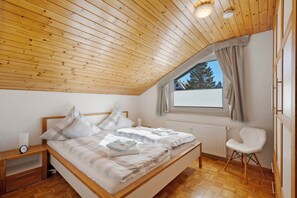 Un dormitorio con techo de madera, cama con ropa blanca, mesita de noche, una silla y ventana con vistas.