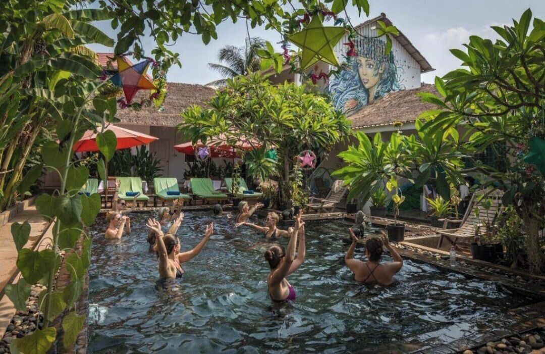 A pool area with people enjoying the water, surrounded by lush greenery and a mural on the building.