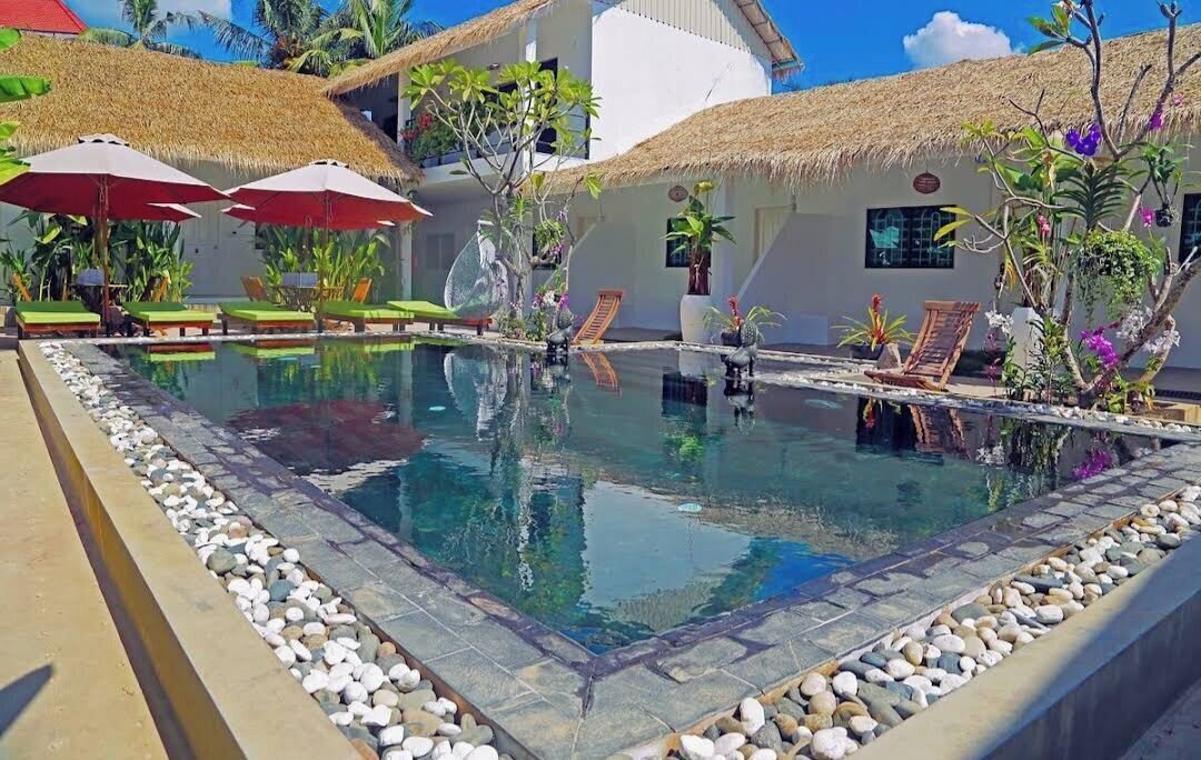 A pool area with lounge chairs and umbrellas, surrounded by tropical plants and a thatched-roof building.