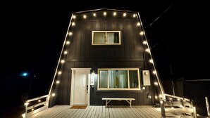 A dark-colored building with a white door and windows, a wooden deck, and string lights.