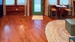 Una cabaña acogedora con paredes de madera, una puerta verde y un ventilador de techo. Hay ventanas con cortinas, una mesa con sillas y una alfombra en el suelo.
