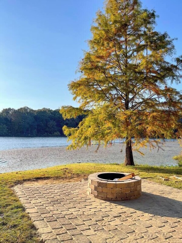 A stone fire pit surrounded by a paved area with a view of a lake and trees in the background.