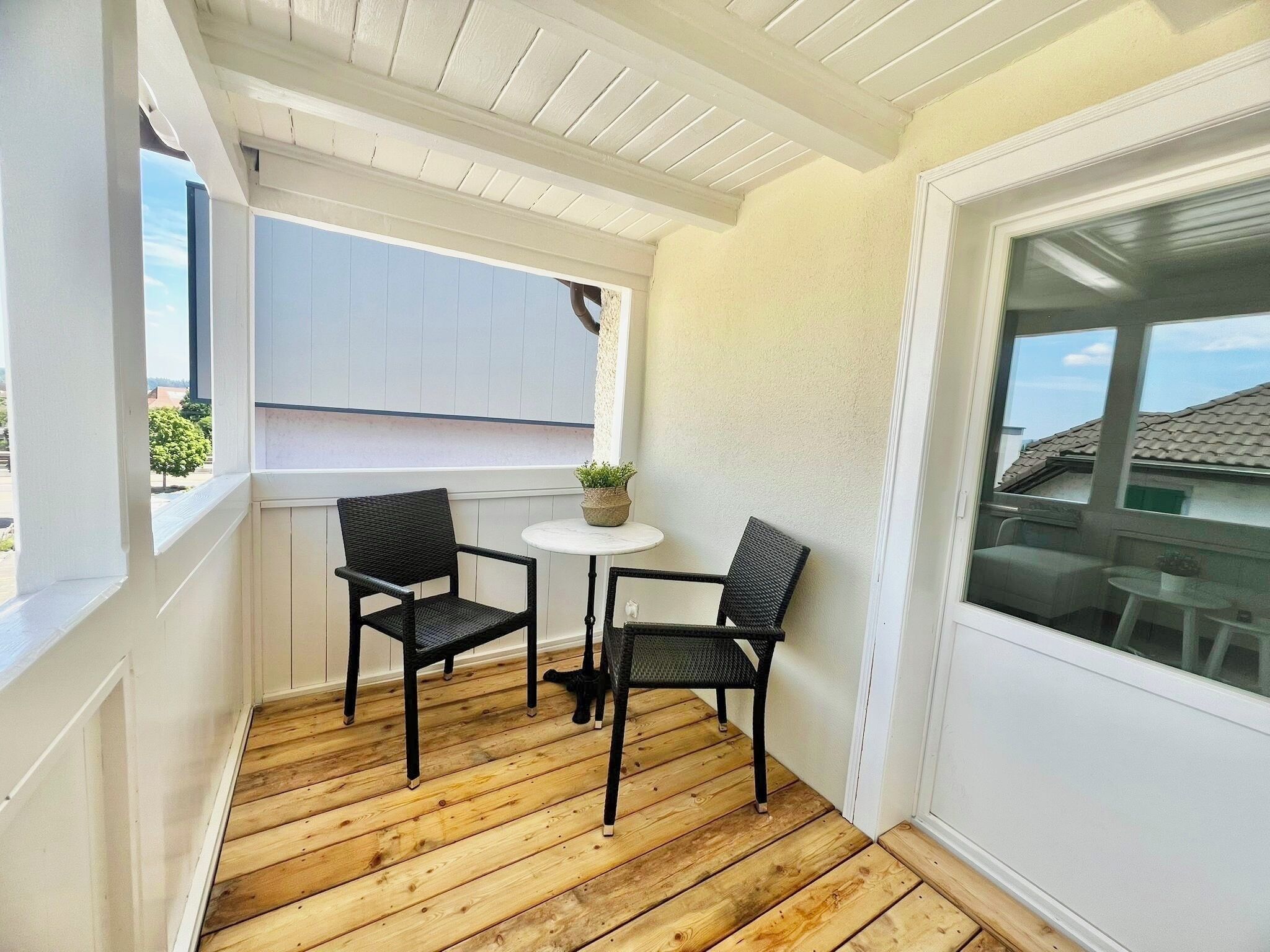 A wooden deck with two wicker chairs and a small round table, a potted plant, and a window with a view of a house.