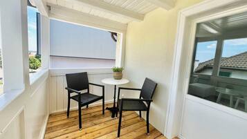A wooden deck with two wicker chairs and a small round table, a potted plant, and a window with a view of a house.