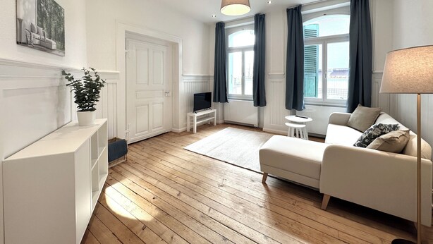 A modern living room with a wooden floor, a white sofa, a small white table, a TV on a stand, a potted plant, and a large window with curtains. - Business Apartment Niederbipp (Niederbipp)