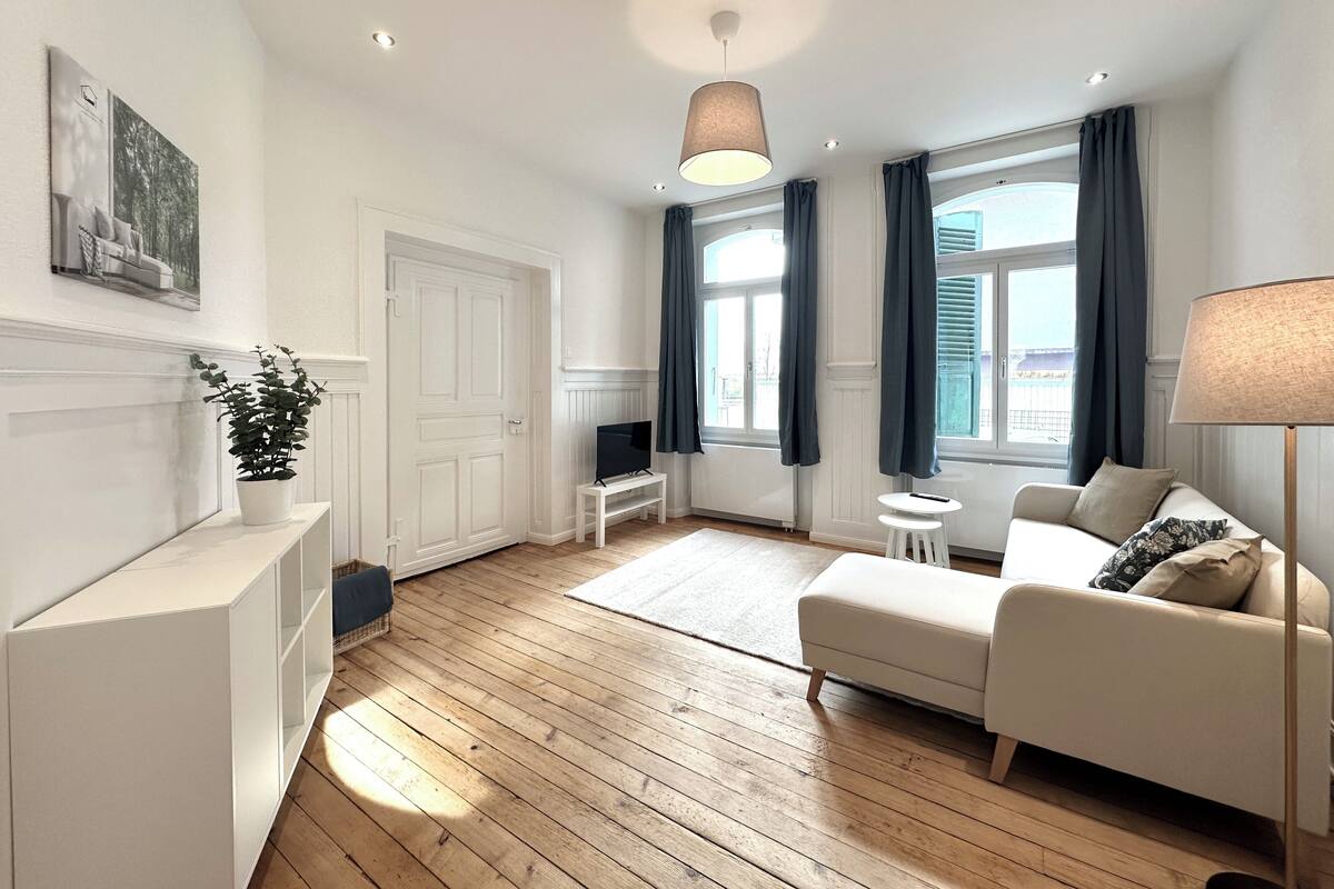 Una sala de estar moderna con piso de madera, un sofá blanco, una mesita pequeña blanca, un televisor sobre un mueble bajo, una planta en maceta y un ventanal con cortinas.