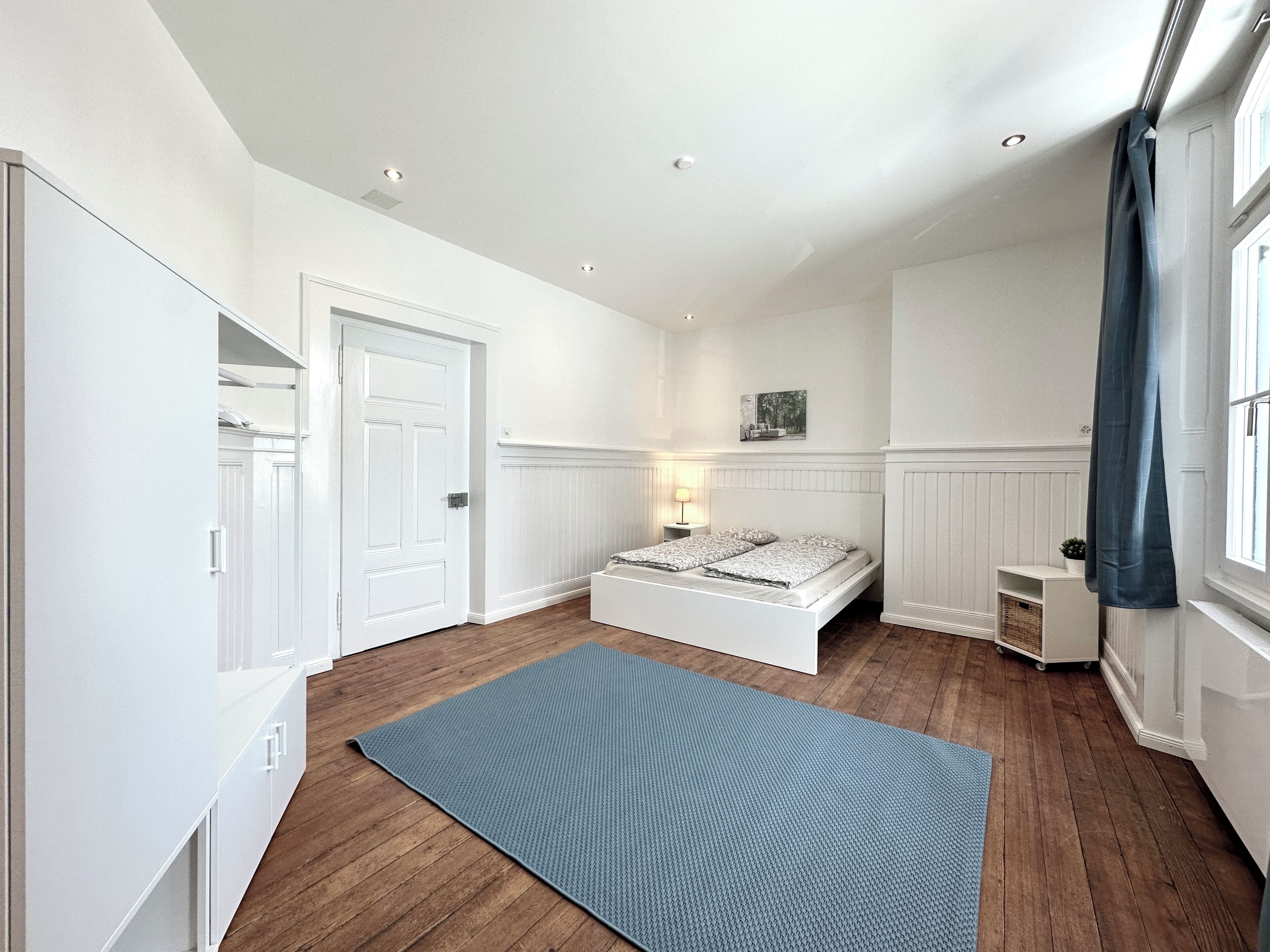Ein Schlafzimmer mit Holzboden, einem Bett mit weißer Bettwäsche, einem Nachttisch, einer Lampe und einem Fenster mit Vorhang.